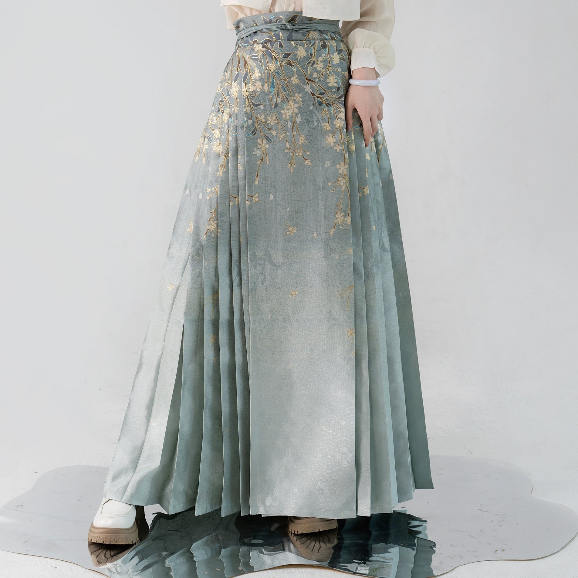 Close-up of pleated Hanfu skirt with gold floral embroidery and gradient green fabric, inspired by Ming-dynasty fashion.