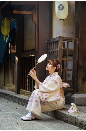 A woman in a beige Japanese kimono dress sitting gracefully with a fan. The yukata features delicate cherry blossom prints.
