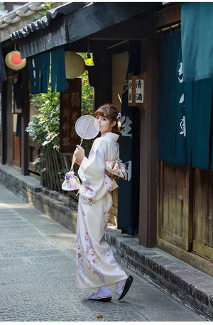 A woman in a Japanese kimono dress holding a fan and matching bag. The beige yukata features cherry blossom prints and an elegant obi.