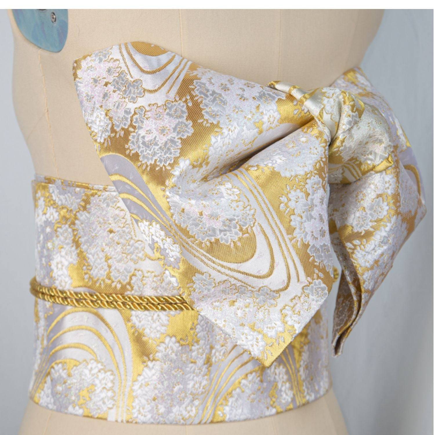 Gold and white brocade bow belt with floral patterns, styled on mannequin for traditional Japanese kimono waist seal and yukata wear.