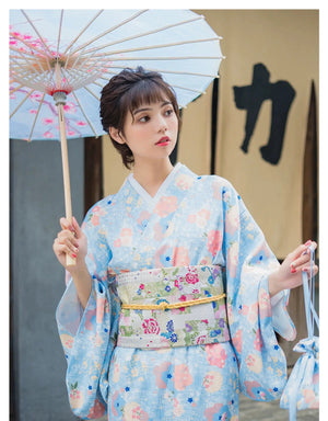 A woman in a light blue floral kimono yukata holding a parasol, styled with a decorative obi. A cute and elegant Japanese-inspired dress.