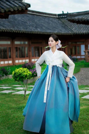 Woman standing near traditional Korean palace in blue hanbok princess dress with white top, floral hair accessory