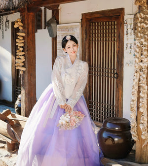 White purple Korean female high-end genuine Yanji court hanbok with floral headpiece, posed near traditional Korean jars and decor.