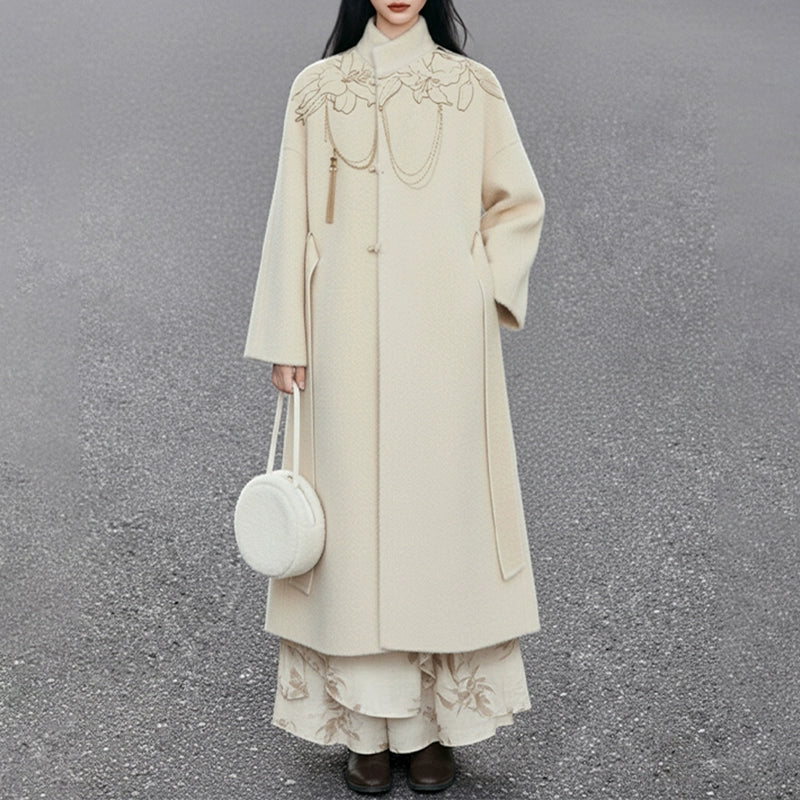 Person wearing a long beige coat with floral embroidery on a gray background