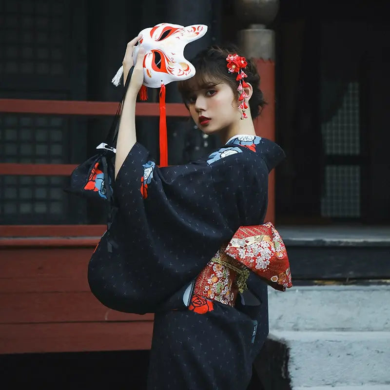 Close-up of a woman in a black Japanese yukata holding a fox mask, showcasing intricate red and blue floral designs. A refined Halloween look.