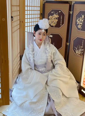 Indoor view of White Yanji Hanbok court dress with embroidered sleeves and lavender sash, traditional Korean attire for daily use.