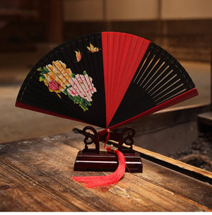 Red and black bamboo fan with floral design on wooden stand. Includes red tassel, perfect for traditional Chinese dance.