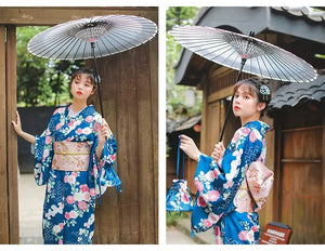 Woman wearing a blue floral kimono with a brocade Yukata bow belt, holding a traditional Japanese umbrella. Elegant styling with a decorative waist seal.