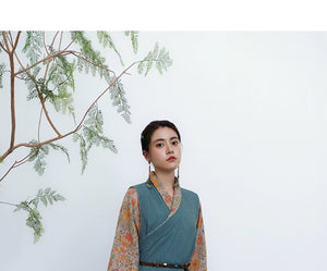 Model wearing teal cross-collared Tibetan top with floral sleeves near decorative branch