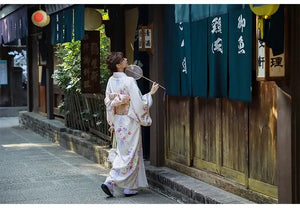 A woman in a Japanese kimono dress walking along a traditional street. The beige yukata features cherry blossom patterns and an obi belt.