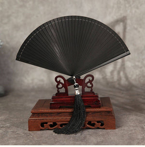 Black bamboo folding fan on wooden stand, minimalist design with black tassel. Suitable for traditional Chinese dance and Hanfu styling.
