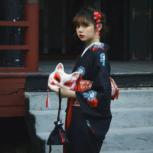 Side view of a woman in a black yukata holding a fox mask and black pouch, adorned with red floral accents. A blend of tradition and elegance.