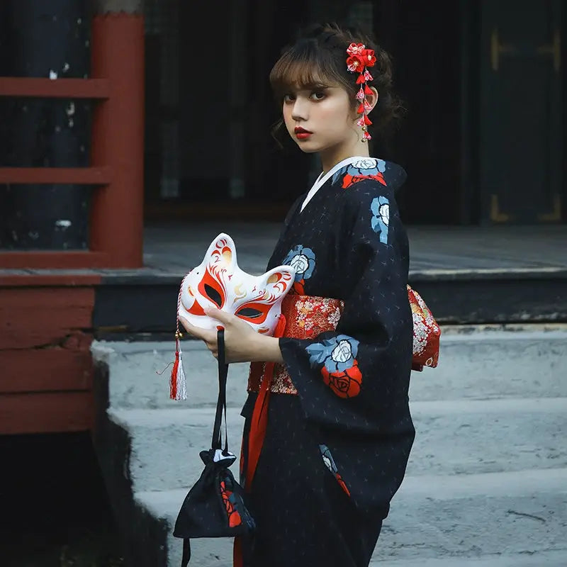 Side view of a woman in a black yukata holding a fox mask and black pouch, adorned with red floral accents. A blend of tradition and elegance.