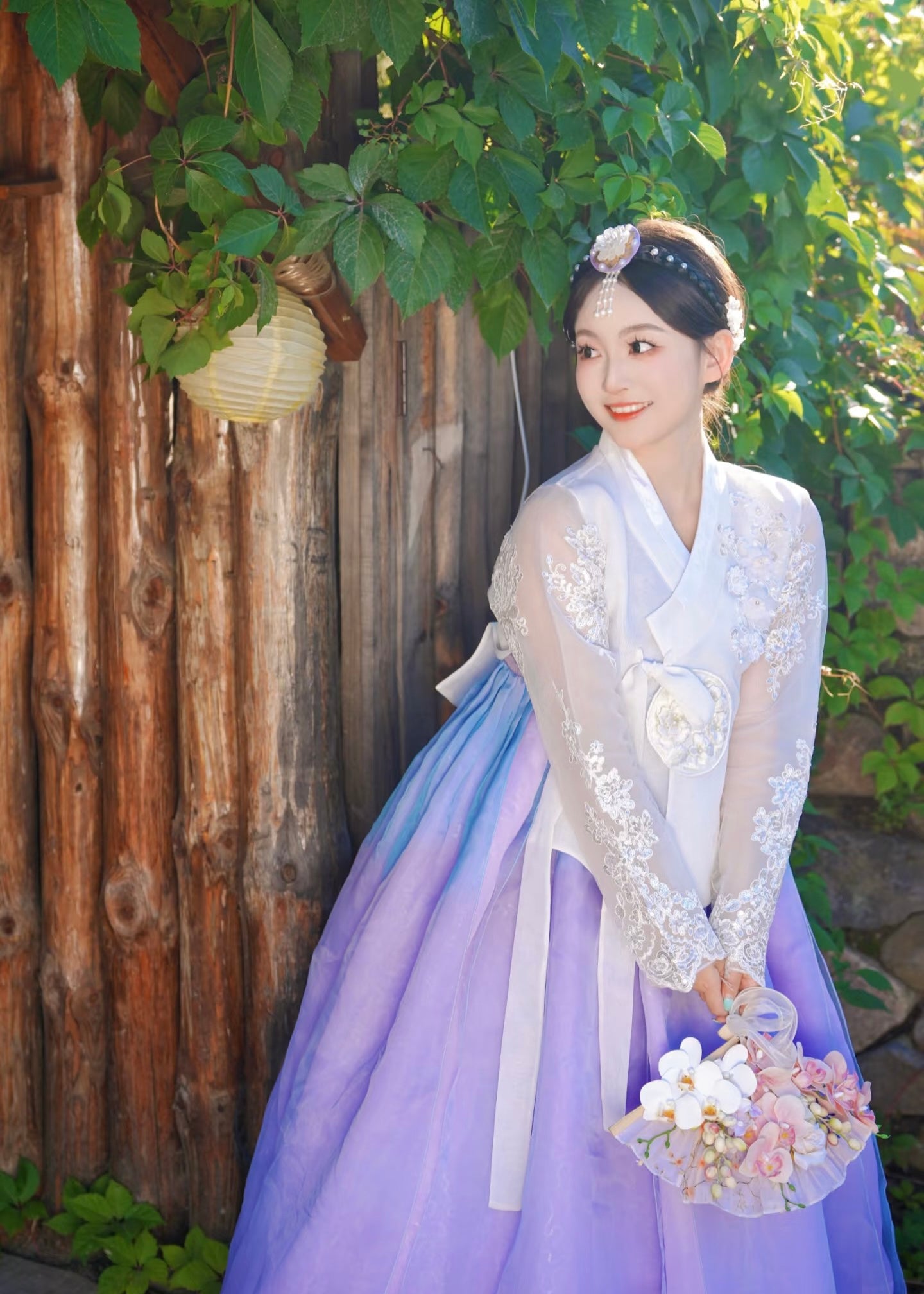 Traditional white purple Korean female high-end genuine Yanji court hanbok with floral bouquet, posed near wooden fence and greenery.
