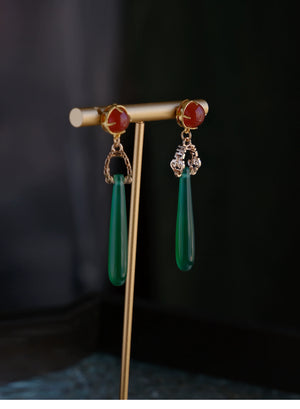 Luxury green agate water drop earrings with red gemstone tops and gold accents, showcased on a modern display stand.