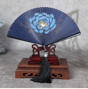 Blue bamboo folding fan with large blue peony flower, displayed on wooden stand. Ideal for Hanfu dance with black tassel.