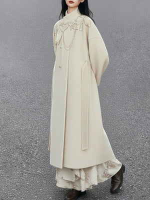 Person wearing a long, light-colored coat with intricate designs on a gray background