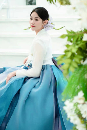 Woman sitting in blue hanbok princess dress with white top, floral hair accessory, near white structure with greenery