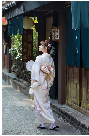 A woman in a Japanese kimono dress walking gracefully, showcasing the obi bow. The beige yukata is adorned with floral patterns.