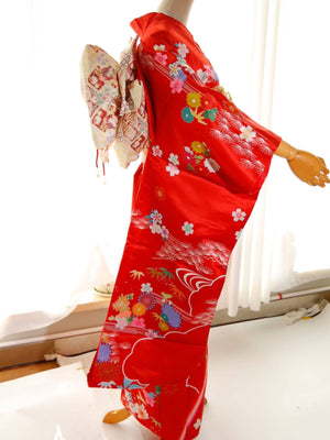 Side view of red swinging sleeve kimono with floral prints and cream-colored big three-leaf bow belt. Geisha-style belt adds traditional charm.