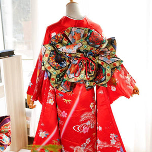 Back view of red kimono with swinging sleeves and large floral-patterned three-leaf bow belt. Traditional geisha-style belt design.
