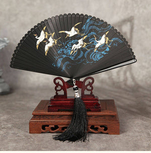 Black folding fan with crane and wave design on wooden stand. Full bamboo fan with black tassel, ideal for Hanfu dance.
