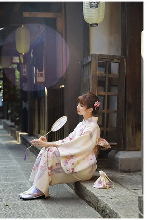 A woman in a Japanese kimono dress gazing into the distance, holding a fan. The beige yukata features cherry blossom designs.