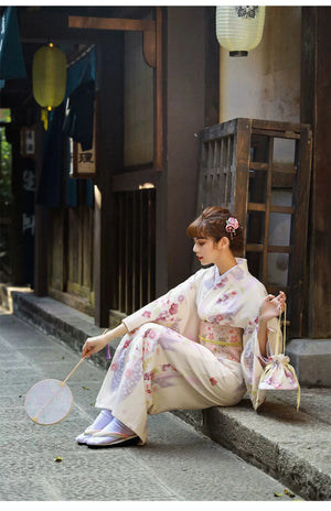 A woman in a Japanese kimono dress sitting elegantly with a fan. The beige yukata is adorned with cherry blossom prints and a matching obi.