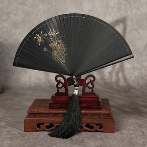 Black bamboo fan with gold floral design on wooden stand. Elegant Hanfu accessory with black tassel for traditional dance.