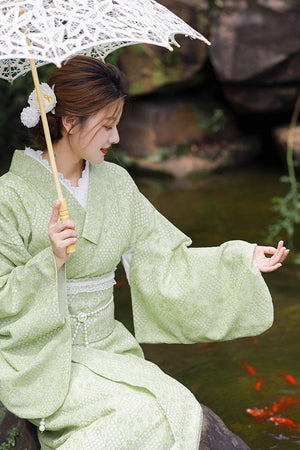 Woman wearing green lace kimono waist seal bow with matching kimono, holding lace parasol beside a koi pond.
