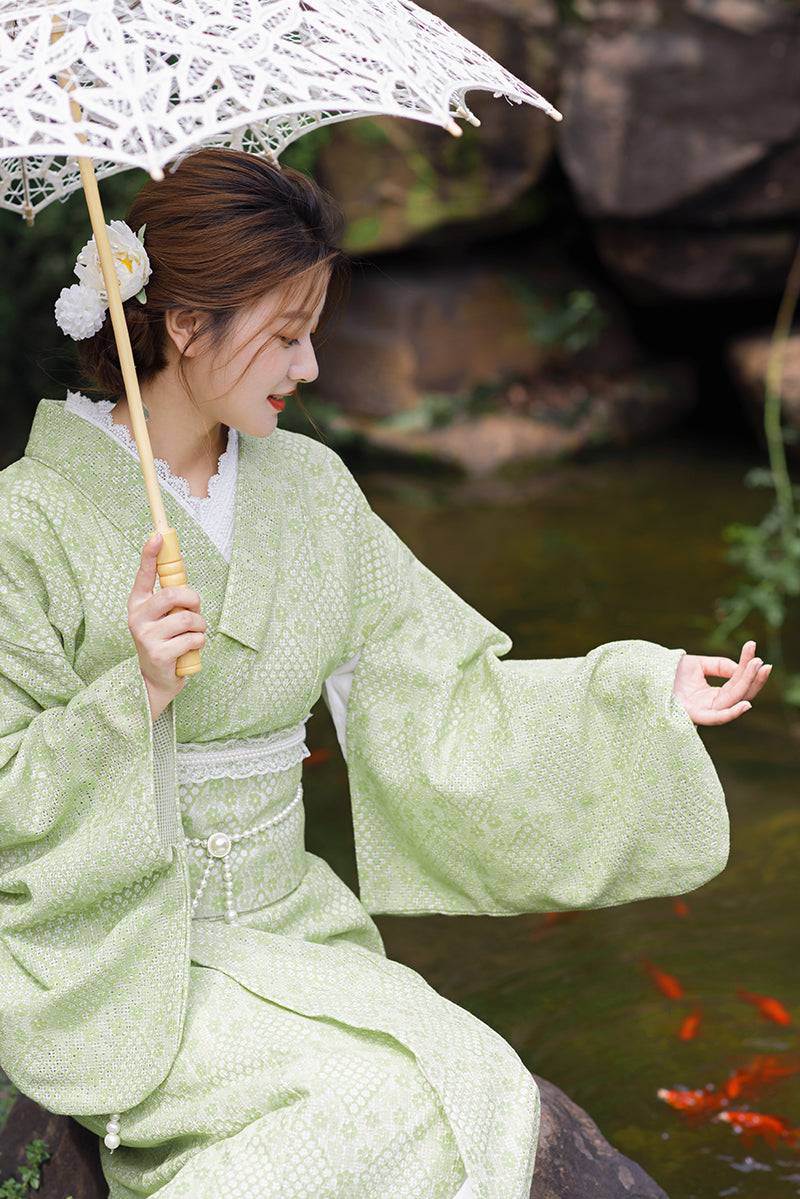 Woman wearing green lace kimono waist seal bow with matching kimono, holding lace parasol beside a koi pond.