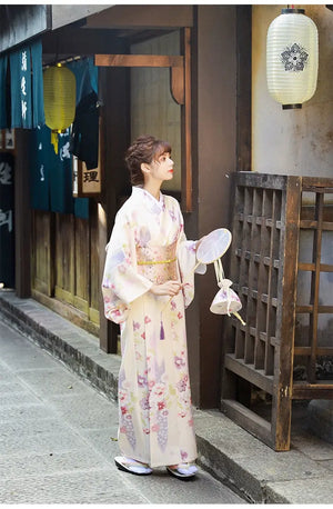 A woman in a Japanese kimono dress standing near a wooden structure, holding a fan. The elegant beige yukata is adorned with floral prints.