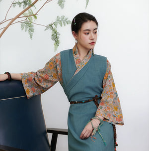 Teal cross-collared Tibetan top with floral sleeves, model leaning on dark furniture against white wall with branch