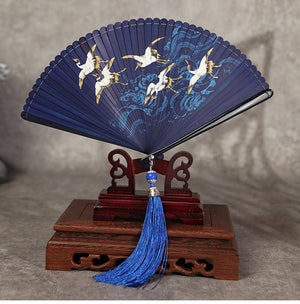 Blue bamboo folding fan with crane and wave design on wooden stand. Features blue tassel, ideal for Hanfu dance.