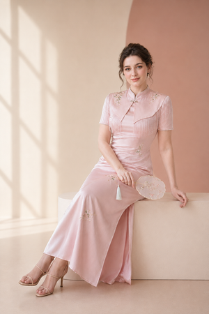 A woman wearing a light pink short-sleeve cheongsam-style dress sits on a beige platform in a softly lit studio. The dress has subtle floral embroidery near the shoulders and hem, and she is holding a matching round fan.