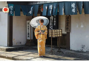 Woman in an orange floral kimono yukata holding a traditional umbrella, standing in front of a Japanese building with wooden decor and banners.