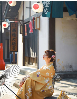 Side view of a woman in an orange floral kimono yukata sitting on a stone pavement, surrounded by traditional Japanese street decor.