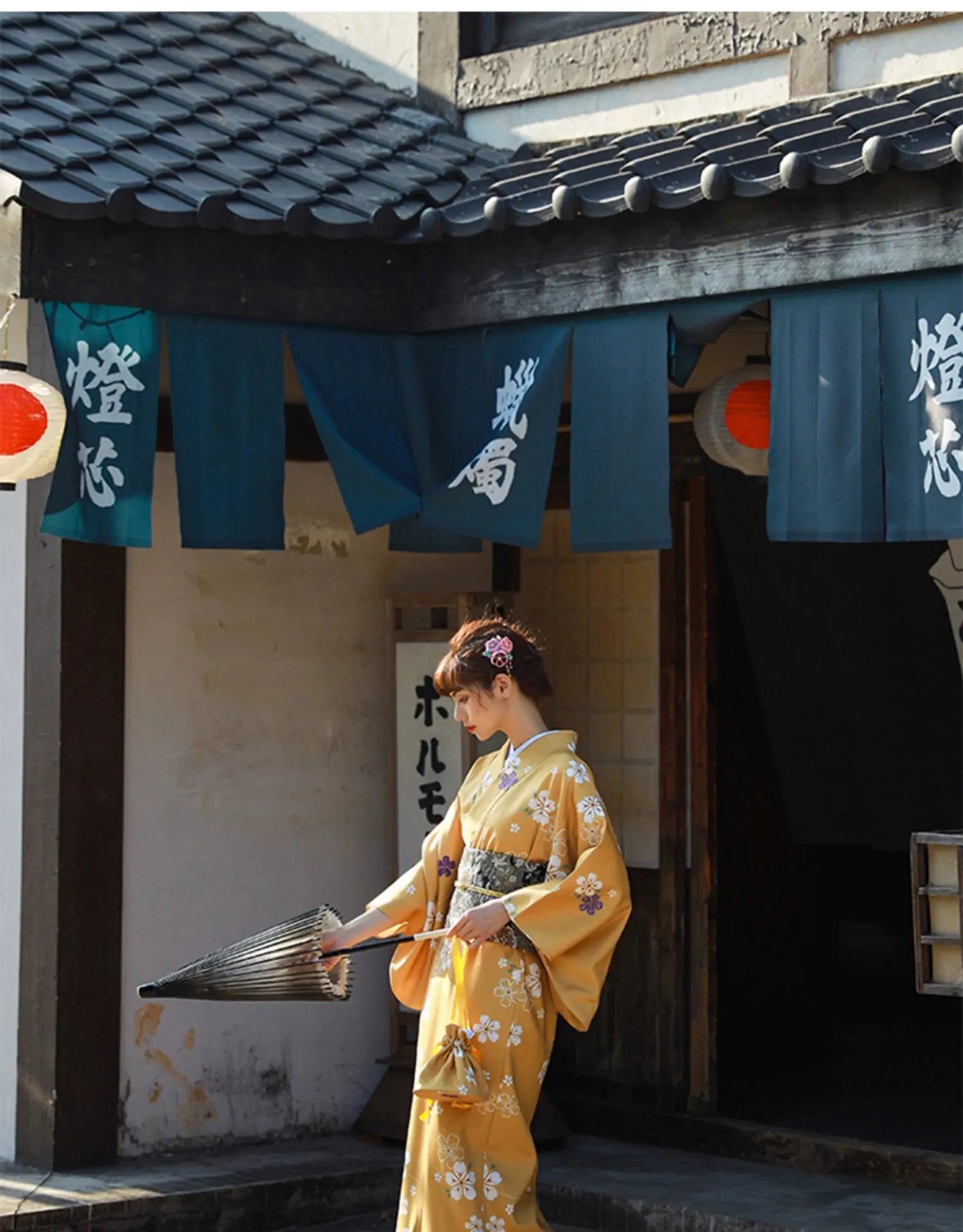 Woman in an orange floral kimono yukata holding a folded umbrella, standing near a traditional Japanese building with blue banners and lanterns.