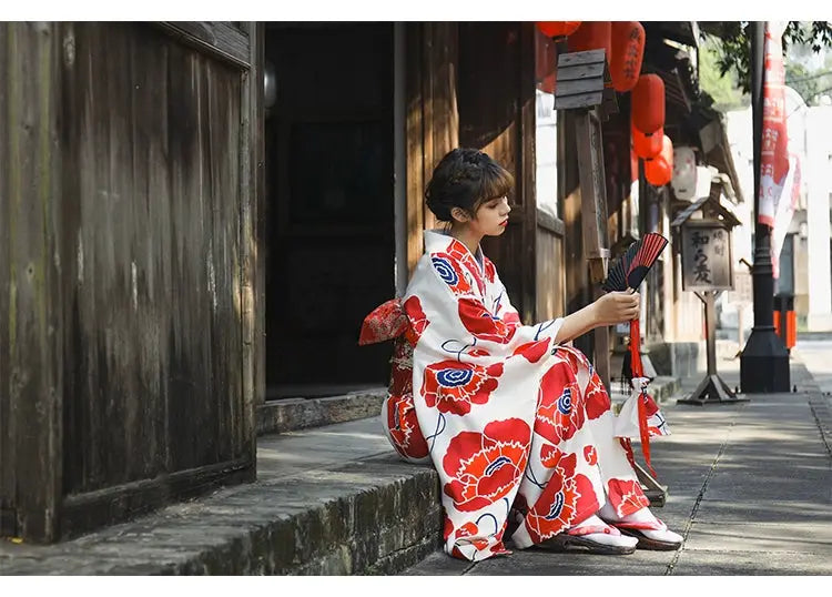 Traditional Japanese kimono dress in white and red floral print, styled with an obi belt, worn by a woman seated on a stone pathway.