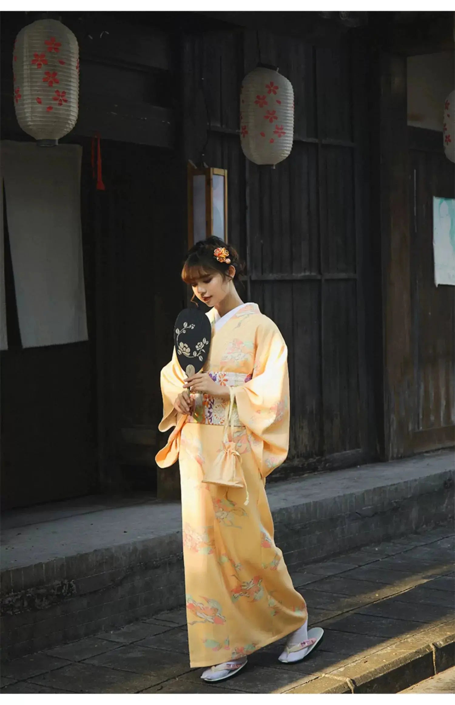 Woman in an orange Japanese kimono-style yukata with floral patterns, holding a decorative fan while walking in a traditional wooden street.