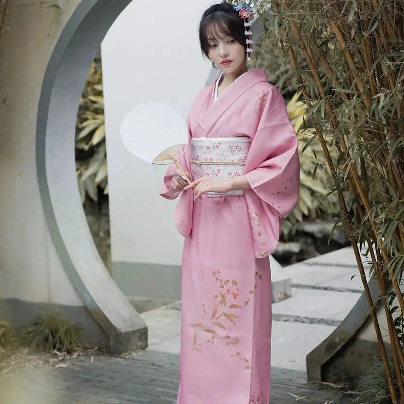 Full-length pink Japanese kimono yukata with floral accents, styled with a white obi, showcased in a serene outdoor setting.