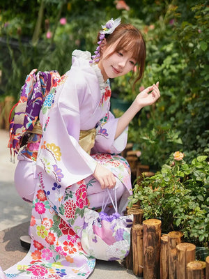 Woman in a pastel floral Japanese kimono kneeling in a garden, showcasing traditional design details and a matching handbag.