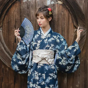 Woman in a blue floral yukata holding a fan, styled with a white brocade kimono belt seal featuring subtle pink and green accents.