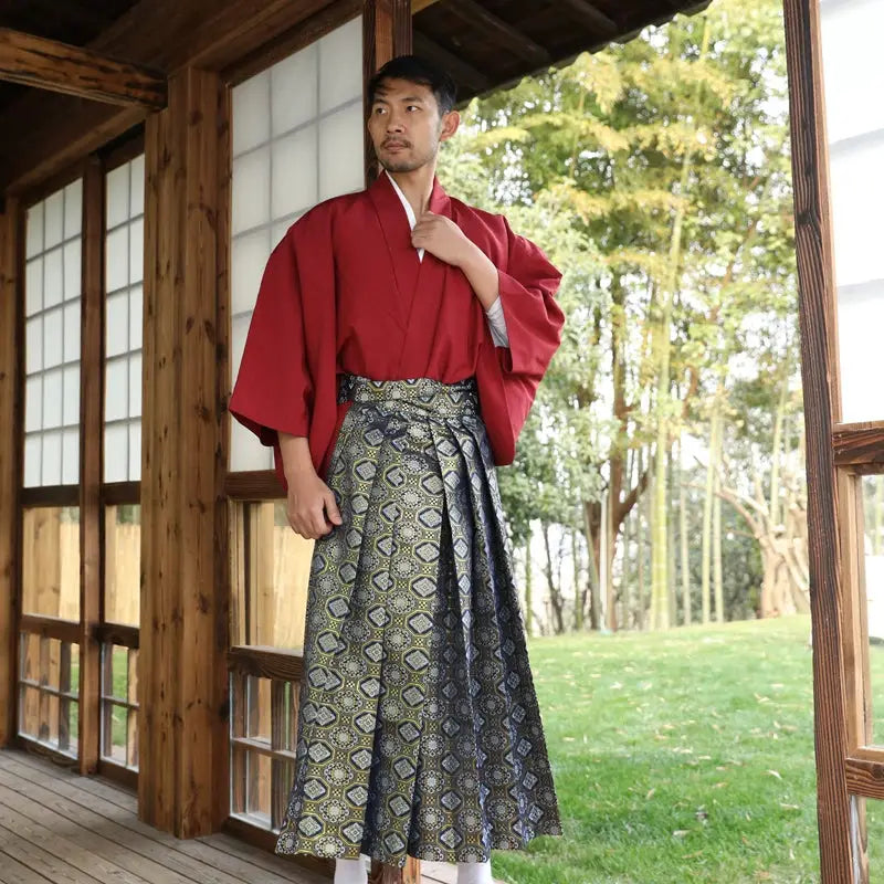 Man dressed in a Japanese traditional kimono with a red top and intricate brocade skirt, posing near a wooden structure with a garden view.