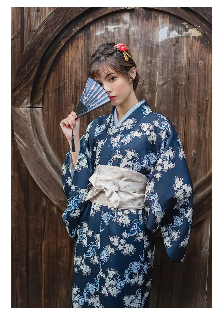 Elegant blue floral yukata paired with a white brocade kimono belt seal, worn by a woman holding a fan in a traditional Japanese setting.