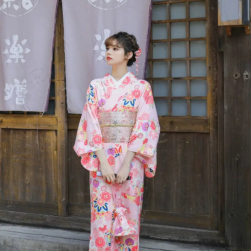 Front view of a woman in a pink floral kimono with a decorative obi belt, standing in front of a traditional Japanese wooden building.