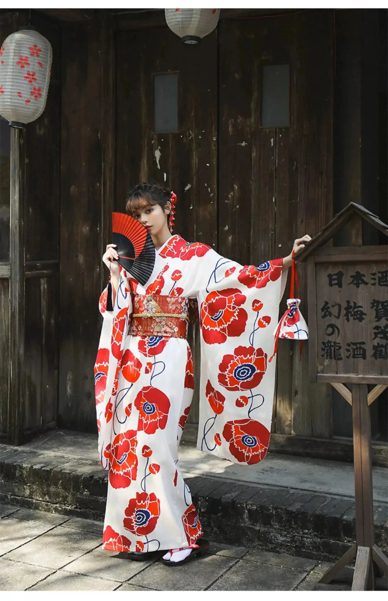 Woman posing in a white and red floral Japanese kimono dress with wide sleeves, holding a fan in front of a wooden building.