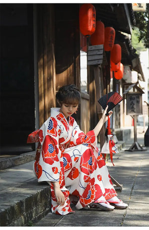 Woman in a white and red floral Japanese kimono dress, seated outdoors holding a fan, showcasing the traditional design and accessories.