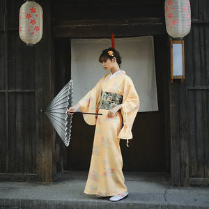 Full-length view of a woman wearing a formal orange Japanese kimono-style yukata with floral designs, holding a black umbrella in front of a traditional wooden building.