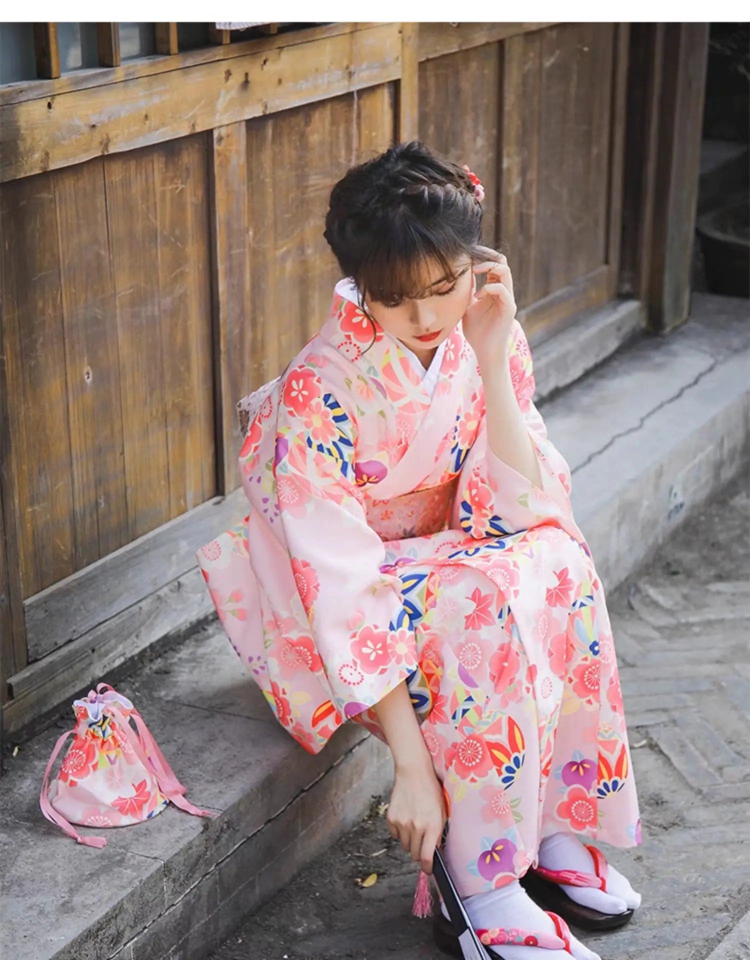 Seated woman in a pink cherry blossom kimono, holding a fan and a matching floral pouch, near a traditional wooden building.